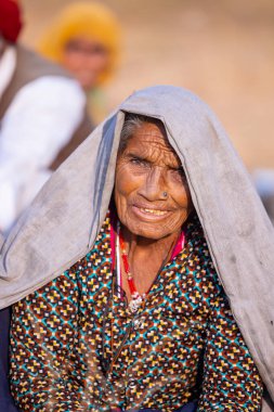 Puşkar, Rajasthan, Hindistan - 24 Kasım 2023: Pushkar Deve Fuarı sırasında yerde oturan renkli geleneksel elbise giymiş yaşlı Hintli Rajasthani kadının portresi.