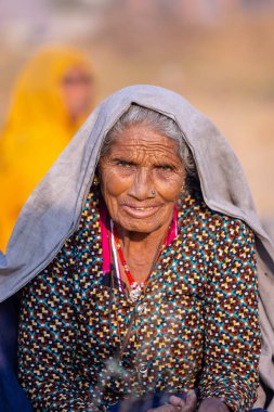 Puşkar, Rajasthan, Hindistan - 24 Kasım 2023: Pushkar Deve Fuarı sırasında yerde oturan renkli geleneksel elbise giymiş yaşlı Hintli Rajasthani kadının portresi.