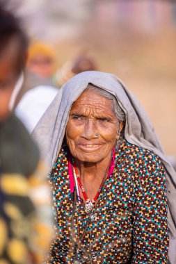 Puşkar, Rajasthan, Hindistan - 24 Kasım 2023: Pushkar Deve Fuarı sırasında yerde oturan renkli geleneksel elbise giymiş yaşlı Hintli Rajasthani kadının portresi.