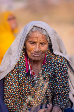 Puşkar, Rajasthan, Hindistan - 24 Kasım 2023: Pushkar Deve Fuarı sırasında yerde oturan renkli geleneksel elbise giymiş yaşlı Hintli Rajasthani kadının portresi.