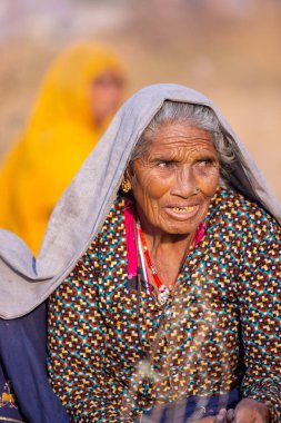 Puşkar, Rajasthan, Hindistan - 24 Kasım 2023: Pushkar Deve Fuarı sırasında yerde oturan renkli geleneksel elbise giymiş yaşlı Hintli Rajasthani kadının portresi.