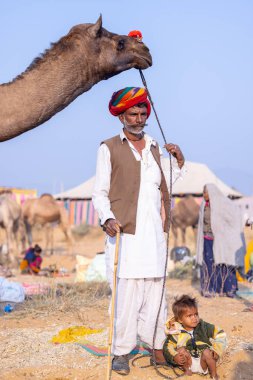 Puşkar, Rajasthan, Hindistan - 23 Kasım 2023: Geleneksel beyaz elbiseli ve renkli türbanlı yaşlı adamın portresi ve Pushkar deve fuarı sırasında devenin yanında duran küçük çocuk. 
