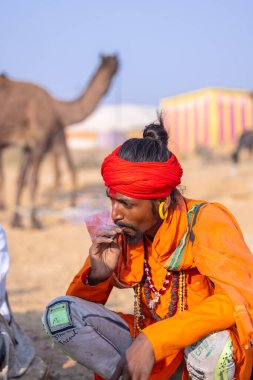 Puşkar, Rajasthan, Hindistan - 24 Kasım 2023: İtme arabası fuarı sırasında açık alanda sigara içen kırmızı türbanlı genç bir erkek yılan portresi. 