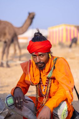 Puşkar, Rajasthan, Hindistan - 24 Kasım 2023: İtme arabası fuarı sırasında açık alanda sigara içen kırmızı türbanlı genç bir erkek yılan portresi. 