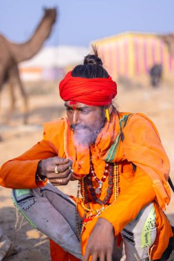 Puşkar, Rajasthan, Hindistan - 24 Kasım 2023: İtme arabası fuarı sırasında açık alanda sigara içen kırmızı türbanlı genç bir erkek yılan portresi. 