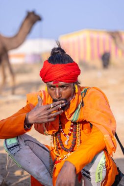 Puşkar, Rajasthan, Hindistan - 24 Kasım 2023: İtme arabası fuarı sırasında açık alanda sigara içen kırmızı türbanlı genç bir erkek yılan portresi. 