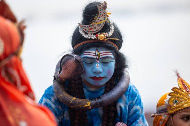 Varanasi, Uttar Pradesh, Hindistan - 20 Ocak 2024: Varanasi 'de kış sabahı Ganga nehrinde boyalı yüzüyle yürüyen Lord Shiv gibi giyinmiş genç bir çocuğun portresi.