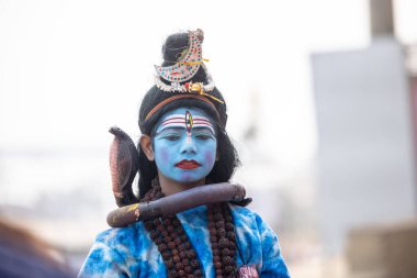 Varanasi, Uttar Pradesh, Hindistan - 20 Ocak 2024: Varanasi 'de kış sabahı Ganga nehrinde boyalı yüzüyle yürüyen Lord Shiv gibi giyinmiş genç bir çocuğun portresi.