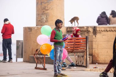 Varanasi, Uttar Pradesh, Hindistan - 20 Ocak 2024: Varanasi 'de kış akşamı kenevir nehrinde boyalı yüzlü uçan uçurtma gibi giyinmiş genç bir çocuğun portresi.
