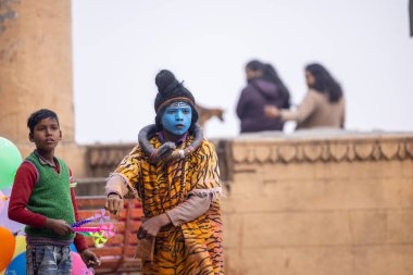 Varanasi, Uttar Pradesh, Hindistan - 20 Ocak 2024: Varanasi 'de kış akşamı kenevir nehrinde boyalı yüzlü uçan uçurtma gibi giyinmiş genç bir çocuğun portresi.
