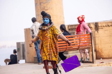 Varanasi, Uttar Pradesh, Hindistan - 20 Ocak 2024: Varanasi 'de kış sabahı Ganga nehrinde boyalı yüzüyle yürüyen Lord Shiv gibi giyinmiş genç bir çocuğun portresi.