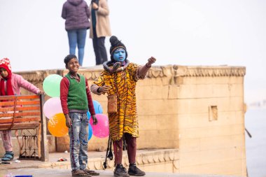Varanasi, Uttar Pradesh, Hindistan - 20 Ocak 2024: Varanasi 'de kış sabahı Ganga nehrinde boyalı yüzüyle yürüyen Lord Shiv gibi giyinmiş genç bir çocuğun portresi.