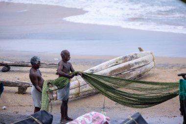 Thiruvananthapuram, Kerala, Hindistan - 14 Ekim 2024: Balıkları balık ağından çıkarmak için sahile park etmiş ahşap teknenin yanında duran yaşlı erkek balıkçılar grubu. İş yerinde tanıştık..