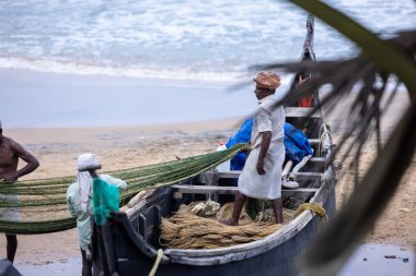 Thiruvananthapuram, Kerala, Hindistan - 14 Ekim 2024: Balıkları balık ağından çıkarmak için sahile park etmiş ahşap teknenin yanında duran yaşlı erkek balıkçılar grubu. İş yerinde tanıştık..