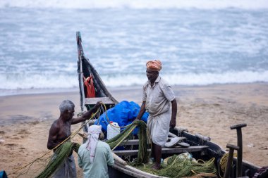 Thiruvananthapuram, Kerala, Hindistan - 14 Ekim 2024: Balıkları balık ağından çıkarmak için sahile park etmiş ahşap teknenin yanında duran yaşlı erkek balıkçılar grubu. İş yerinde tanıştık..
