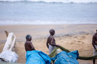Thiruvananthapuram, Kerala, Hindistan - 14 Ekim 2024: Balıkları balık ağından çıkarmak için sahile park etmiş ahşap teknenin yanında duran yaşlı erkek balıkçılar grubu. İş yerinde tanıştık..
