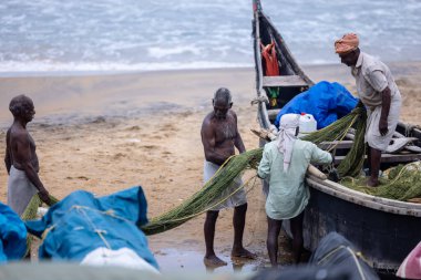 Thiruvananthapuram, Kerala, Hindistan - 14 Ekim 2024: Balıkları balık ağından çıkarmak için sahile park etmiş ahşap teknenin yanında duran yaşlı erkek balıkçılar grubu. İş yerinde tanıştık..