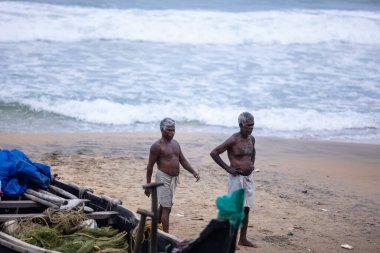 Thiruvananthapuram, Kerala, Hindistan - 14 Ekim 2024: Balıkları balık ağından çıkarmak için sahile park etmiş ahşap teknenin yanında duran yaşlı erkek balıkçılar grubu. İş yerinde tanıştık..