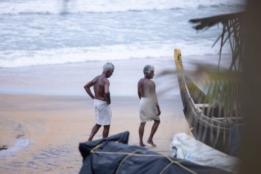 Thiruvananthapuram, Kerala, Hindistan - 14 Ekim 2024: Balıkları balık ağından çıkarmak için sahile park etmiş ahşap teknenin yanında duran yaşlı erkek balıkçılar grubu. İş yerinde tanıştık..