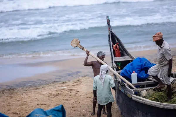 Thiruvananthapuram, Kerala, Hindistan - 14 Ekim 2024: Balıkları balık ağından çıkarmak için sahile park etmiş ahşap teknenin yanında duran yaşlı erkek balıkçılar grubu. İş yerinde tanıştık..