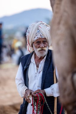 Puşkar, Rajasthan, Hindistan - Kasım 09 2024: Geleneksel beyaz giysili rajasthani erkek portresi çöl panayırında deve fuarı sırasında deve ticareti sırasında sarıklı.