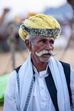 Puşkar, Rajasthan, Hindistan - Kasım 09 2024: Geleneksel beyaz giysili rajasthani erkek portresi çöl panayırında deve fuarı sırasında deve ticareti sırasında sarıklı.