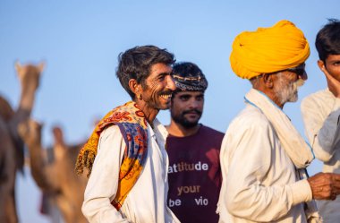 Puşkar, Rajasthan, Hindistan - Kasım 09 2024: Geleneksel beyaz elbiseli bir rajasthani erkeğinin portresi çöl panayırında deve pazarında deve ticareti yaparken renkli sarıklı.
