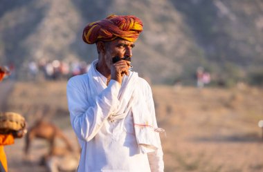 Puşkar, Rajasthan, Hindistan - Kasım 09 2024: Geleneksel beyaz elbiseli bir rajasthani erkeğinin portresi çöl panayırında deve pazarında deve ticareti yaparken renkli sarıklı.
