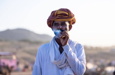 Puşkar, Rajasthan, Hindistan - Kasım 09 2024: Geleneksel beyaz elbiseli bir rajasthani erkeğinin portresi çöl panayırında deve pazarında deve ticareti yaparken renkli sarıklı.
