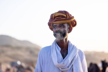 Puşkar, Rajasthan, Hindistan - Kasım 09 2024: Geleneksel beyaz elbiseli bir rajasthani erkeğinin portresi çöl panayırında deve pazarında deve ticareti yaparken renkli sarıklı.