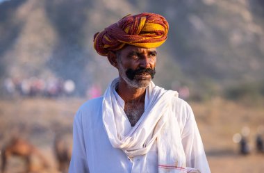 Puşkar, Rajasthan, Hindistan - Kasım 09 2024: Geleneksel beyaz elbiseli bir rajasthani erkeğinin portresi çöl panayırında deve pazarında deve ticareti yaparken renkli sarıklı.
