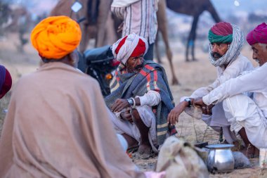 Puşkar, Rajasthan, Hindistan - Kasım 09 2024: Geleneksel beyaz elbiseli bir rajasthani erkeğinin portresi çöl panayırında deve pazarında deve ticareti yaparken renkli sarıklı.