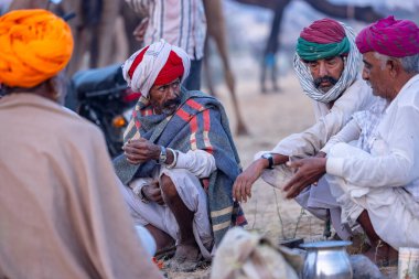 Puşkar, Rajasthan, Hindistan - Kasım 09 2024: Geleneksel beyaz elbiseli bir rajasthani erkeğinin portresi çöl panayırında deve pazarında deve ticareti yaparken renkli sarıklı.