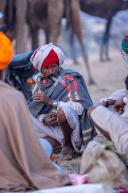 Puşkar, Rajasthan, Hindistan - Kasım 09 2024: Geleneksel beyaz elbiseli bir rajasthani erkeğinin portresi çöl panayırında deve pazarında deve ticareti yaparken renkli sarıklı.
