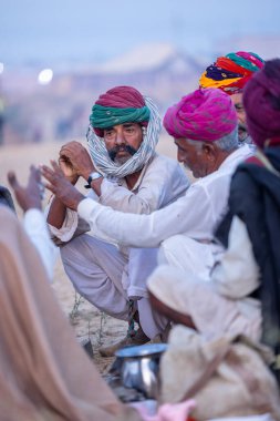Puşkar, Rajasthan, Hindistan - Kasım 09 2024: Geleneksel beyaz elbiseli bir rajasthani erkeğinin portresi çöl panayırında deve pazarında deve ticareti yaparken renkli sarıklı.