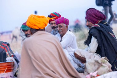 Puşkar, Rajasthan, Hindistan - Kasım 09 2024: Geleneksel beyaz elbiseli bir rajasthani erkeğinin portresi çöl panayırında deve pazarında deve ticareti yaparken renkli sarıklı.