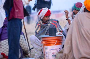 Puşkar, Rajasthan, Hindistan - Kasım 09 2024: Geleneksel beyaz elbiseli bir rajasthani erkeğinin portresi çöl panayırında deve pazarında deve ticareti yaparken renkli sarıklı.
