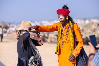 Pushkar, Rajasthan, Hindistan - 10 Kasım 2024: İtme kar deve fuarı sırasında açık zeminde renkli türbanlı kobra yılanı tutan genç yılan oynatıcısı erkek portresi.