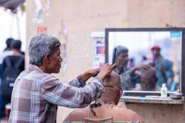 Varanasi, Uttar Pradesh, Hindistan - 20 Ocak 2024: Varanasi 'de Hindu ayinlerinin bir parçası olarak Dashashwamedh ghat' ta yaşlı bir erkeğe tıraş olan bir erkek sokak berberinin portresi.