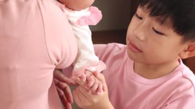 Brother boy is excited with the newborn girl and want to play with an infant while the mother hold toddler in arm, Asian sibling play together in the bedroom having fun and cheerful, smiling boy looks to younger sister and kiss with love and care