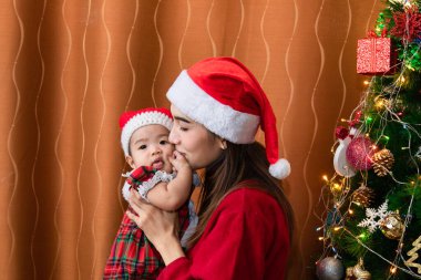 Asyalı mutlu bir anne ve süveter giyen sevimli bir bebek Noel ağacı dekorasyonuna yakın otururlar. Tatil zamanı evde birlikte oynarlar. Noel Baba 'nın kırmızı şapkalı güzel annesi tatlı kızı öper.