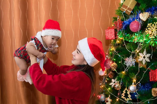 Asian happy young mother and adorable baby on sweater clothes sit near Christmas tree decoration play together at home in Holiday greeting season, beautiful mom with Santa red hat kissing sweet girl