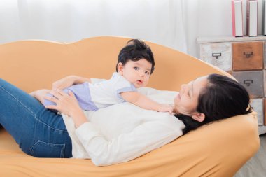 Seçici odak noktası sağlıklı Asyalı anne yeni doğmuş bebeği oturma odasında kucaklıyor, annesi kahverengi kanepeye neşeli ve şefkatli bir şekilde oturuyor. Sevimli küçük çocuk evde annesiyle eğleniyor..