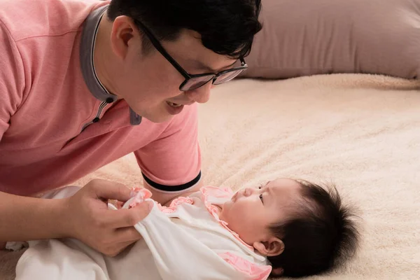 Asian father sing or talk with adorable daughter on bed, close up dad eye contact with healthy newborn 3 months with love and happy, toddler infant lie down on bed look to dad with curiosity.