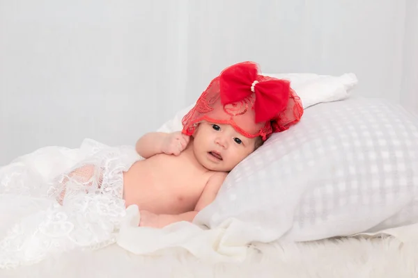 亚洲新生儿身穿红色花边帽服装 看着相机 蹒跚学步的婴儿躺在家里的一张白色床上 — 图库照片