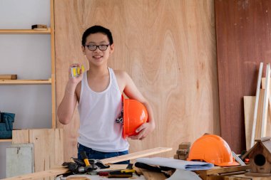 Lovely Asian carpenter boy acting present his work in front of DIY woodworking table carpentry shop with copy space, adorable Chinese boy smiling hold equipment for carpenter tool in workshop at home