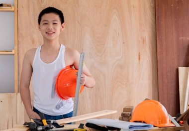 Lovely Asian carpenter boy acting present his work in front of DIY woodworking table carpentry shop with copy space, adorable Chinese boy smiling hold equipment for carpenter tool in workshop at home