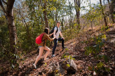 Young Asian tourists couple climbing on rock, man helping hand women hiking looking view beautiful nature outdoor. Partner or couple adventure travel together in forest summer stand on peak mountain
