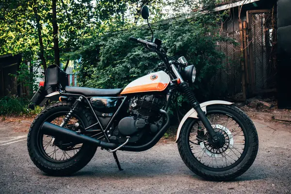 stock image Editorial use only. A vintage-style Suzuki motorcycle with an orange and white color scheme is parked on a quiet street in Kyiv, Ukraine surrounded by lush green trees and foliage.