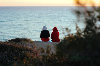 Güneş batarken okyanusa bakan bir uçurumda oturan iki kişinin yakın çekimi. Gün batımı manzarası, okyanus ufku, açık hava rahatlaması, doğa kaçışı, sakin bir an, sakin bir kıyı şeridi, manzaralı bir manzara..